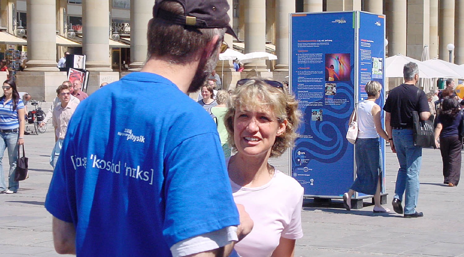 Spiel der Kräfte: viereckiger Plakataufsteller und Ausstellungsmitarbeiter im blauen Event-Shirt "Fragen kostet nichts" in der Fußgängerzone von Stuttgart